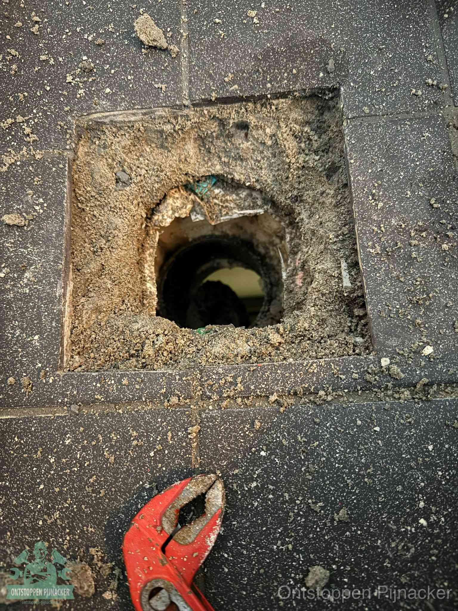 Een geopend rioolputje in een betontegelvloer met losgemaakt rooster en een zichtbare ronde afvoerbuis, het achterhalen waar de afvoerverstopping zit in Pijnacker