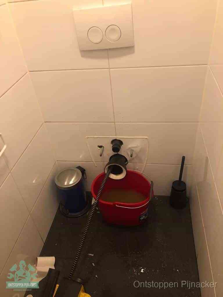 Een ontstopte wand-wc met verwijderde toiletpot waar een ontstoppingsveer in de wanduitgang verdwijnt, het achterhalen waar de verstopping zit in Pijnacker