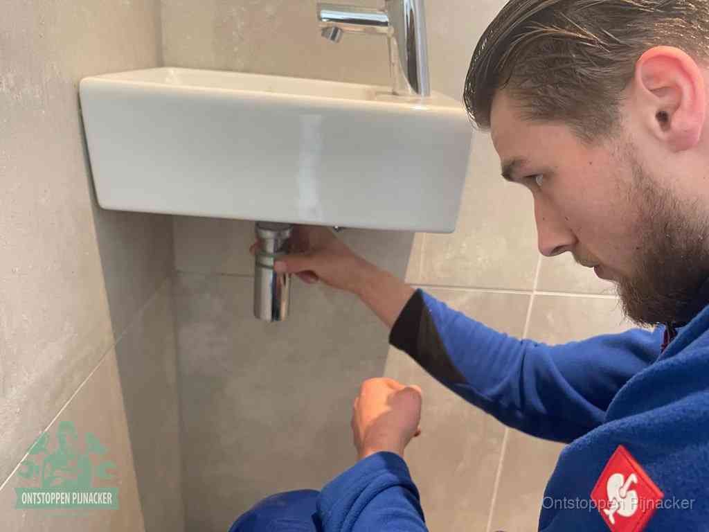 Een monteur draait met de hand de zwanenhalssifon onder een fonteinwasbak in de toiletruimte los om de bron van rioollucht in Pijnacker te vinden
