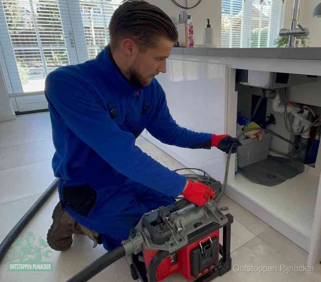 Een monteur met rode handschoenen knielt in een keuken en voert een elektrische ontstoppingsveer in tijdens het professioneel afvoer ontstoppen in Pijnacker